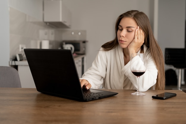 Porträt einer jungen Frau, die vor einem Laptop sitzt und gähnt. Schönes Mädchen in einem weißen Bademantel gekleidet