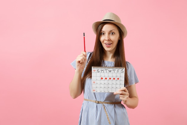 Porträt einer jungen Frau in blauem Kleid, Hut mit rotem Bleistift, Kalender für weibliche Perioden zur Überprüfung der Menstruationstage einzeln auf rosafarbenem Hintergrund. Medizinisches Gesundheitswesen, gynäkologisches Konzept. Platz kopieren