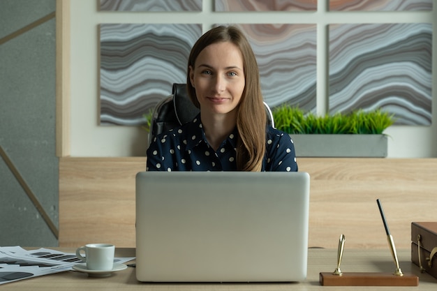 Porträt einer jungen Frau in einem Hemd, die im Büro an einem Laptop arbeitet und in die Kamera schaut