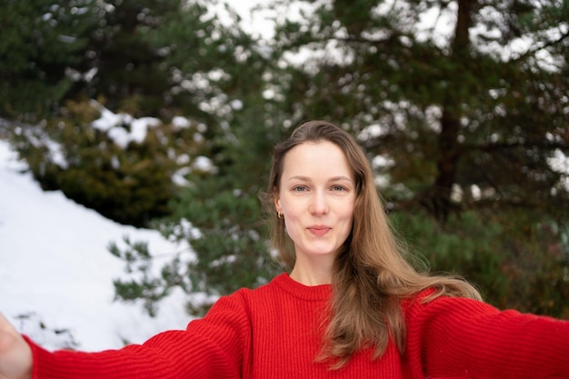 Porträt einer jungen Frau in einem Pullover, die vor dem Hintergrund von Weihnachtsbäumen und Schnee ein Selfie macht oder bei einem Videoanruf spricht. Konzept der Winterferien
