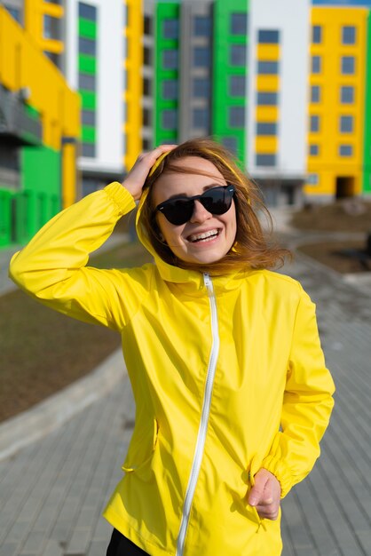 Porträt einer jungen Frau in einer gelben Jacke mit Sonnenbrille, die mit hellen Gebäuden im Hintergrund lächelt