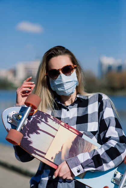 Porträt einer jungen Frau in einer medizinischen Maske mit Longboard in der Stadt während der Quarantäne