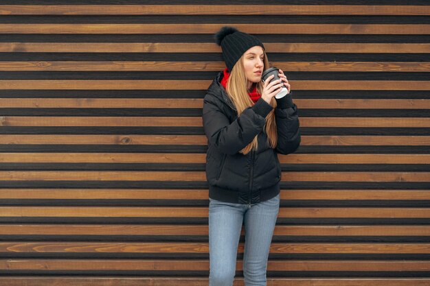 Porträt einer jungen Frau in Winterkleidung, die in einer Straße gegen Holz steht und Kaffee trinkt