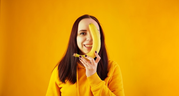 Porträt einer jungen Frau mit Banane auf gelbem Hintergrund Lustige Frau im gelben Hoodie, die mit Früchten posiert