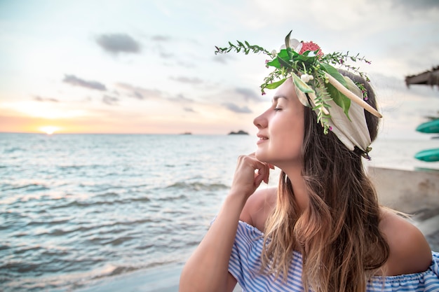 Porträt einer jungen Frau mit einer Zusammensetzung der Blumen auf ihrem Kopf gegen den Ozean bei Sonnenuntergang.