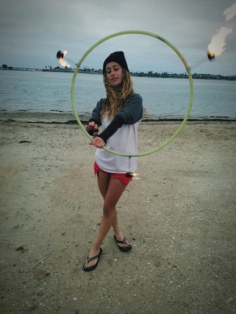 Porträt einer jungen Frau mit Feuer-Hula-Hoop am Strand gegen den Himmel.