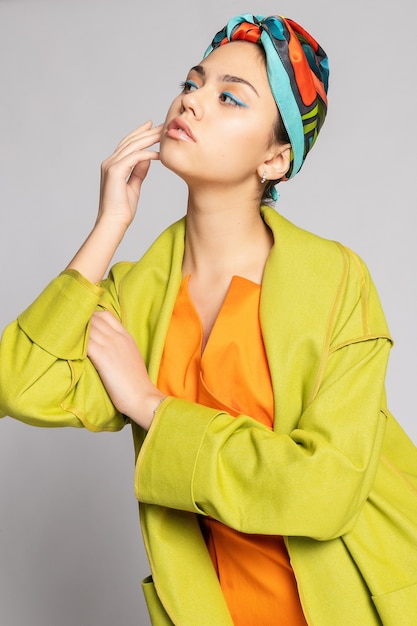 Porträt einer jungen Frau mit hellem Make-up und modischem Kopftuch. Heller Hintergrund. Schönheit, Mode, Make-up-Konzept. Mädchen in einem hellgrünen Mantel, leuchtend orangefarbenem Kleid.