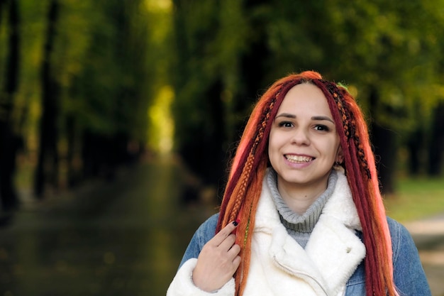Porträt einer jungen Frau mit Ingwer-Dreadlocks, die im Waldpark posieren Hübsche Frau mit heller Frisur in warmer Jeansjacke auf dem Spaziergang