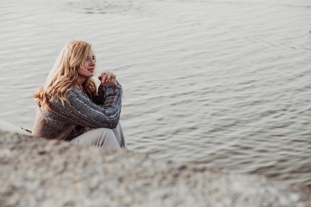 Foto porträt einer jungen frau mit langem, gewelltem, blondem haar, die eine beige hose mit brauner strickjacke trägt, die am ufer des flusses sitzt