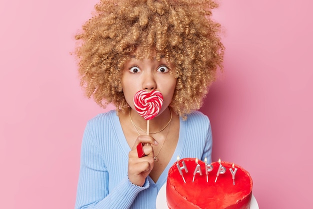 Porträt einer jungen Frau mit lockigem Haar hat sich gewundert, dass ihr Ausdruck den Mund mit herzförmigen Karamellbonbons bedeckt, die köstlichen Kuchen halten, süße Zähne haben und Zuckersucht tragen blaue Pullover-Posen im Innenbereich