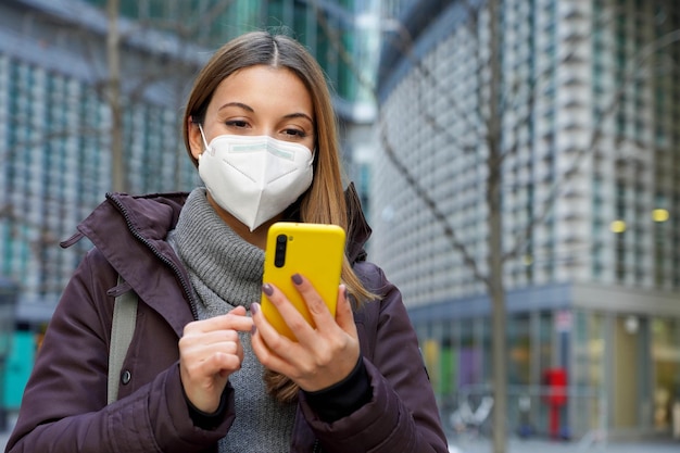 Porträt einer jungen Frau mit medizinischer Gesichtsmaske, die Benachrichtigungen auf dem Smartphone im städtischen Hintergrund überprüft Kopierbereich