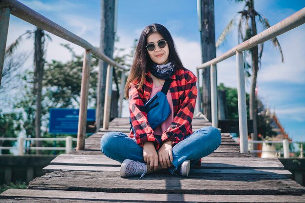 Porträt einer jungen Frau mit Sonnenbrille, die auf einer Fußgängerbrücke gegen den Himmel sitzt