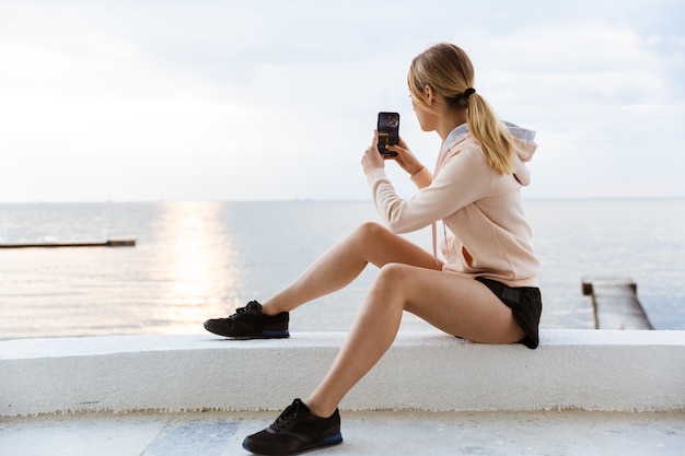 Porträt einer jungen Frau mit Trainingsanzug, die morgens auf dem Pier in der Nähe des Meeres ein Foto auf dem Handy macht