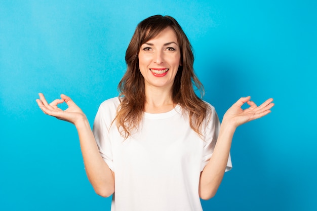 Porträt einer jungen freundlichen Frau im lässigen T-Shirt mit Geste der Meditation und des Lächelns auf Blau. Emotionales Gesicht. Geste zum Entspannen