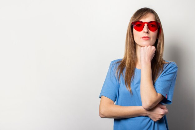 Porträt einer jungen freundlichen Frau in einem lässigen blauen T-Shirt, rote Brille, hält ihre Hand auf ihrem Kinn auf Licht. Emotionales Gesicht. Geste zu denken, zu planen