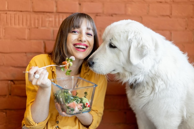 Porträt einer jungen, fröhlichen Frau, die mit ihrem süßen weißen Hund sitzt und gesunden Salat isst