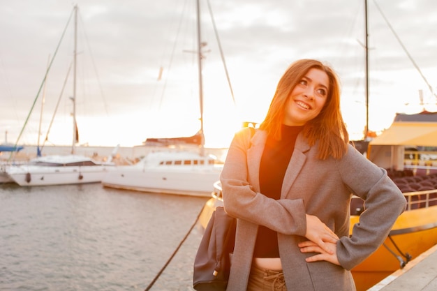 Porträt einer jungen, fröhlichen Frau im Herbstmantel, die bei Sonnenaufgang in einem Yachtclub posiert