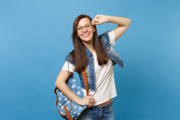 Porträt einer jungen fröhlichen, lustigen, intelligenten Studentin im weißen T-Shirt, Denim-Kleidung mit Rucksack mit Brille stehen isoliert auf blauem Hintergrund. Bildung im Hochschulkonzept der High School.