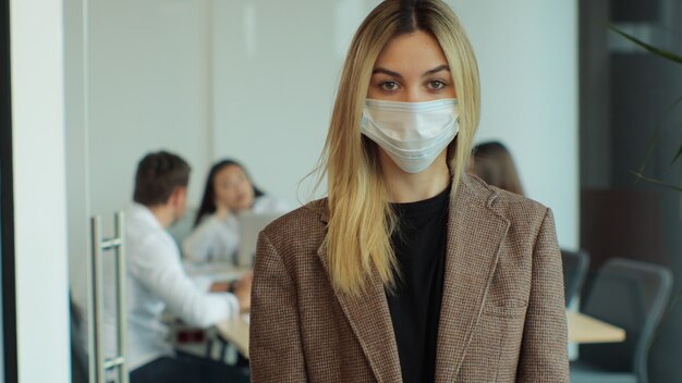 Foto porträt einer jungen geschäftsfrau, die eine schützende gesichtsmaske trägt, die in einem modernen büro steht und die kamera selbstbewusste unternehmerin in sicherheitsmaske betrachtet, die im open space-büro vor der kamera posiert