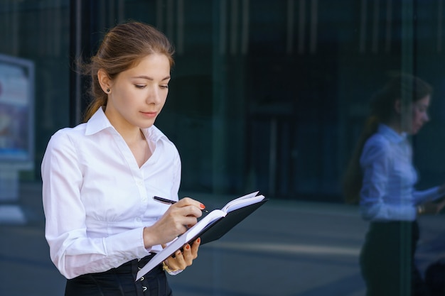 Porträt einer jungen Geschäftsfrau in einem weißen Hemd schreibt in ein Notizbuch nahe einem Bürogebäude