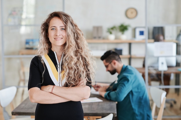 Porträt einer jungen Geschäftsfrau mit langen lockigen Haaren, die mit verschränkten Armen steht und in die Kamera lächelt, während sie mit ihren Kollegen im Büro arbeitet