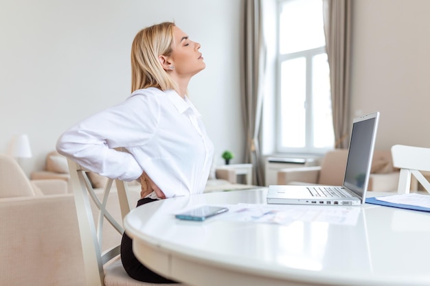 Porträt einer jungen, gestressten Frau, die an ihrem Büro-Schreibtisch vor dem Laptop sitzt, sich den Rücken mit schmerzhaftem Gesichtsausdruck berührt und nach der Arbeit am PC an Rückenschmerzen leidet