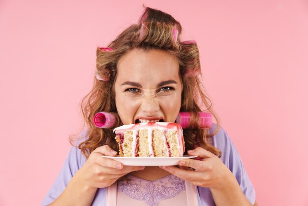 Porträt einer jungen glücklichen Frau in Lockenwicklern, die Kuchen isst, isoliert auf Rosa