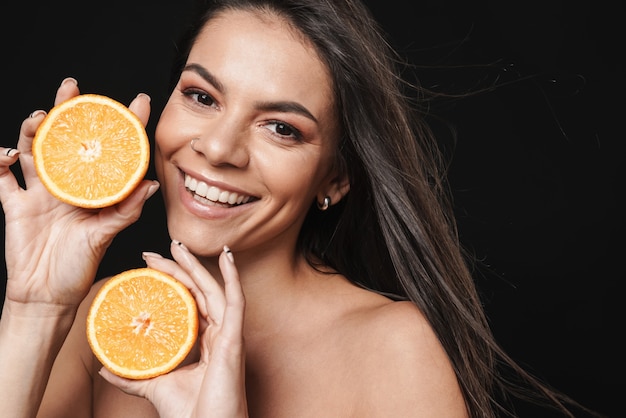 Porträt einer jungen glücklichen schönen nackten Frauenaufstellung lokalisiert über der schwarzen Wand, die orange Zitrusfrucht hält.