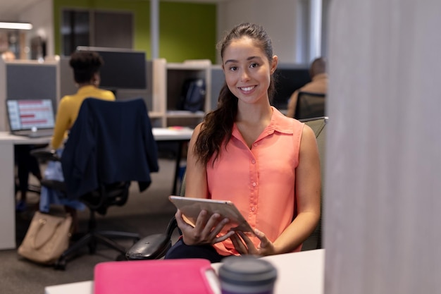 Porträt einer jungen hispanischen Geschäftsfrau, die mit einem digitalen Tablet am modernen Arbeitsplatz sitzt