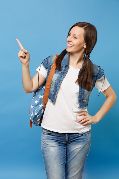 Porträt einer jungen hübschen, fröhlichen Studentin im T-Shirt, Denim-Kleidung mit Rucksack, der den Zeigefinger beiseite zeigt, isoliert auf blauem Hintergrund. Bildung im Gymnasium. Kopieren Sie Platz für Werbung.