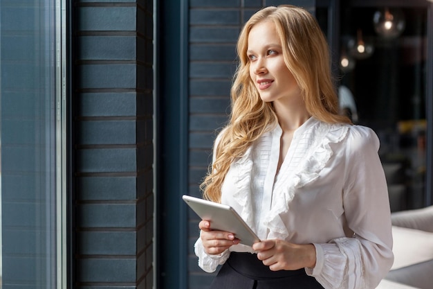 Porträt einer jungen hübschen Geschäftsfrau mit blonden Haaren in stilvollem klassischem Hemd, die aus dem Fenster blickt und lächelnd ein digitales Tablet-PC-Gerät für Büroarbeits-Geschäftsanwendungen im Innenbereich hält