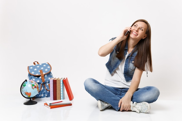 Porträt einer jungen hübschen lachenden Studentin, die auf dem Handy spricht und in der Nähe von Globus sitzt, Rucksack, Schulbücher isoliert