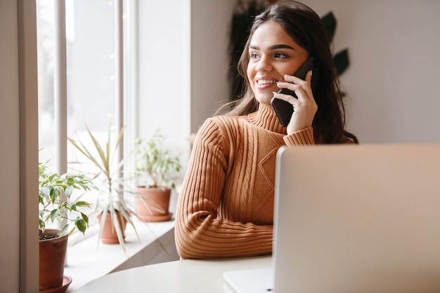 Porträt einer jungen hübschen schönen Frau, die im Café drinnen unter Verwendung des Laptop-Computers sitzt, der durch Handy spricht.