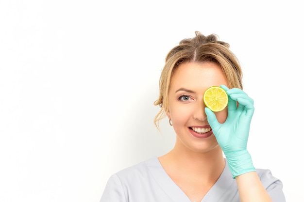 Porträt einer jungen kaukasischen lächelnden Kosmetikerin, die das Auge mit einer Limettenscheibe bedeckt, die Handschuhe trägt