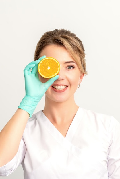 Porträt einer jungen kaukasischen lächelnden Kosmetikerin, die das Auge mit einer Orangenscheibe bedeckt, die Handschuhe über einem weißen Hintergrund trägt