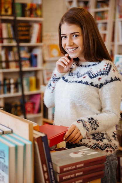 Porträt einer jungen lächelnden Frau, die in der Buchhandlung nach einem Buch sucht und in die Kamera schaut