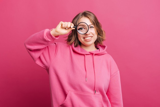 Porträt einer jungen lächelnden Frau im rosa Hoodie, die durch die Lupe schaut