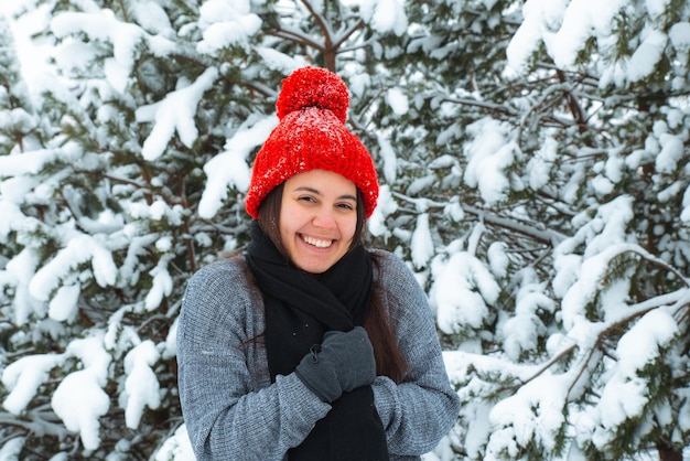 Porträt einer jungen lächelnden Frau in Winterkleidung mit rotem Hut und Bubo-Tanne im Hintergrund. stylisches Konzept