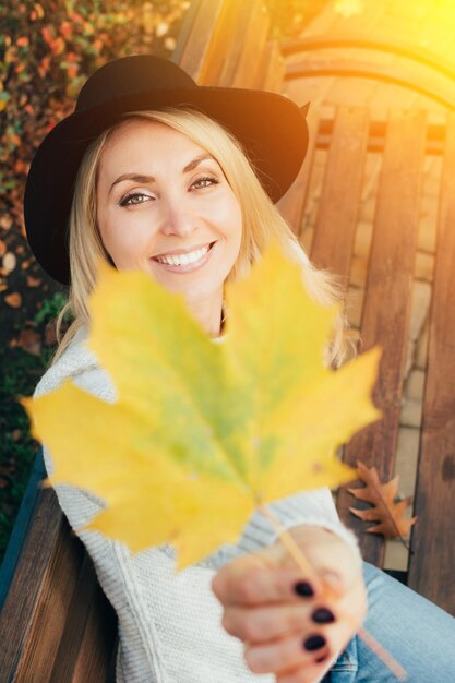 Foto porträt einer jungen lächelnden frau mit einem gelben ahornblatt gegen ihr gesicht mit pullover und hut