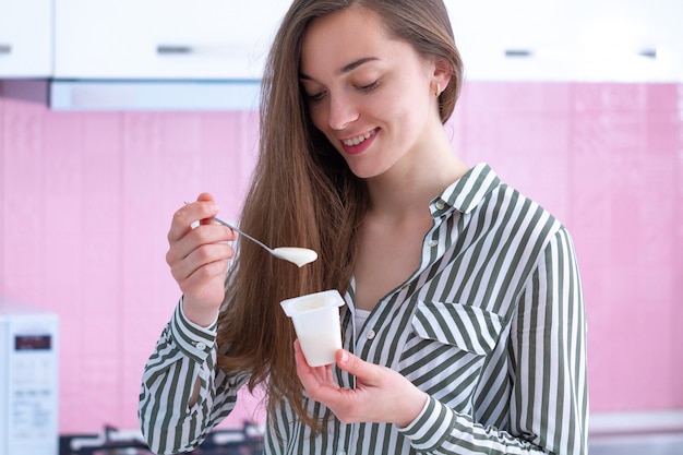 Porträt einer jungen, lächelnden, glücklichen Frau, die Joghurtlöffel hält und zu Hause Joghurt in der Küche isst