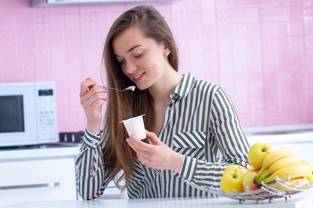 Porträt einer jungen, lächelnden, glücklichen Frau, die zu Hause Joghurt zum Frühstück in der Küche isst