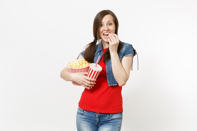 Porträt einer jungen lächelnden hübschen Frau in Freizeitkleidung, die Filmfilm sieht, Popcorn aus dem Eimer isst und Plastikbecher mit Soda oder Cola auf weißem Hintergrund hält. Emotionen im Kinokonzept.