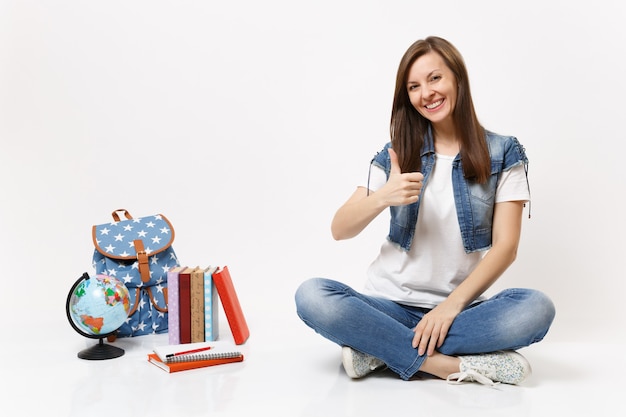Porträt einer jungen, lässig lächelnden Studentin in Denim-Kleidung, die den Daumen zeigt, der in der Nähe von Globus sitzt, Rucksack, Schulbücher isoliert