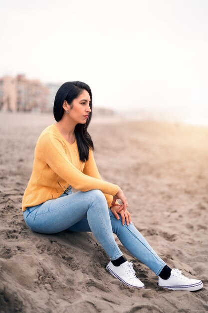 Porträt einer jungen lateinamerikanischen Frau, die am Strand sitzt und den Horizont über dem Ozean betrachtet