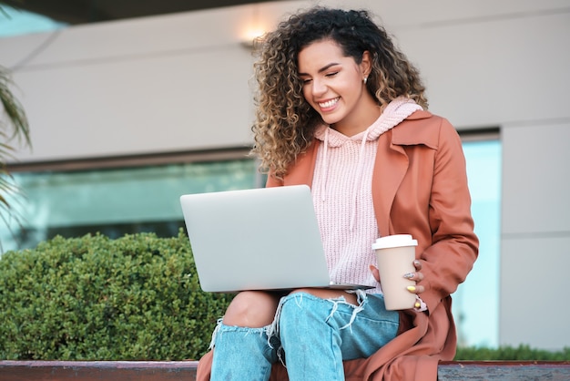 Porträt einer jungen lateinischen Frau, die ihren Laptop verwendet und Kaffee trinkt, während sie draußen sitzt. Urbanes Konzept.
