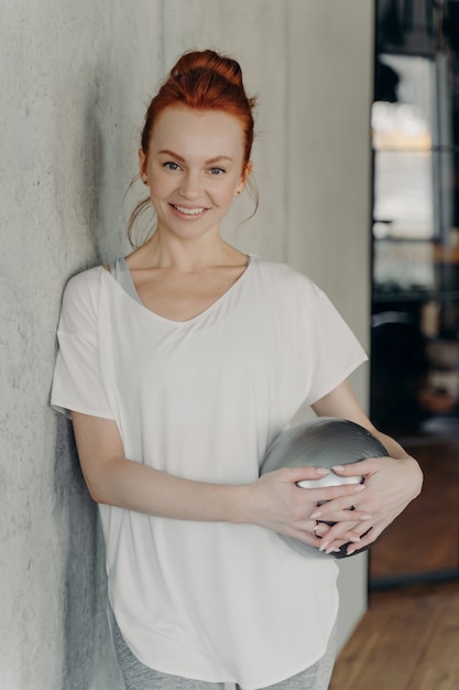 Porträt einer jungen motivierten sportlichen rothaarigen Fitnessfrau, die im Fitnessstudio mit kleinem Gymnastikball steht, sich an eine Betonwand lehnt und glücklich in die Kamera schaut, weißes T-Shirt und graue Leggings trägt