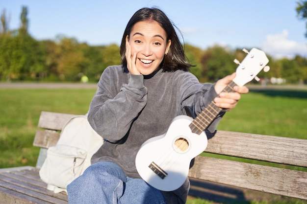 Porträt einer jungen Musikerin, die mit Ukulele-Gitarre im Park sitzt und überrascht auf die Kamera Sayi schaut