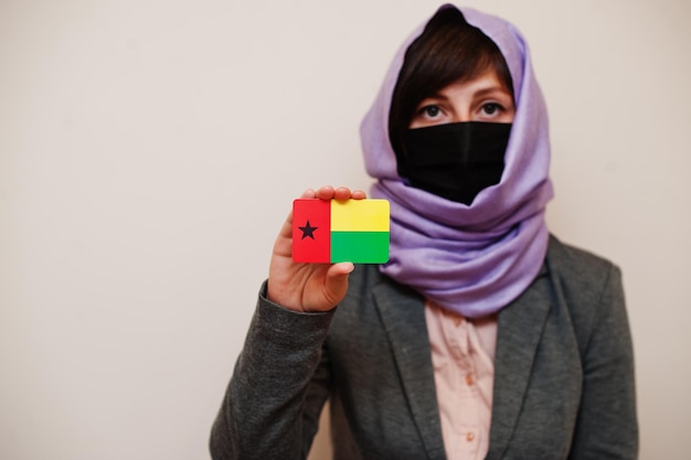 Porträt einer jungen muslimischen Frau, die formelle Kleidung trägt, schützt Gesichtsmaske und Hijab-Kopftuch, hält die Flaggenkarte von Guinea-Bissau vor isoliertem Hintergrund Coronavirus Country Concept