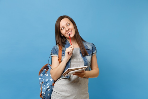 Porträt einer jungen nachdenklichen Studentin in Denim-Kleidung mit Rucksack, die träumendes Denken mit Notizbuch, Bleistift einzeln auf blauem Hintergrund nachschlägt. Ausbildung am Gymnasium.