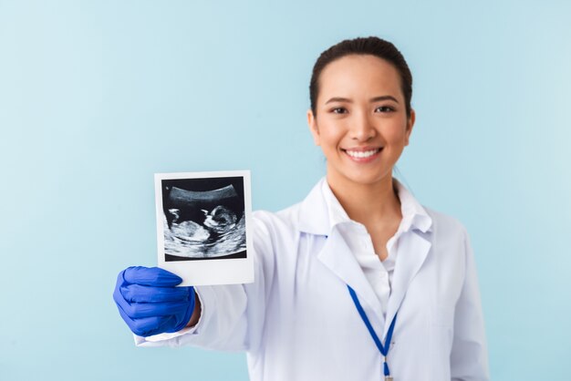 Porträt einer jungen Ärztin, die isoliert über der blauen Wand posiert, die Röntgenaufnahme des Babys in den Händen hält.